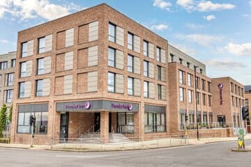 Cozy hotel room with soft beige bedding, a modern white lamp on a wooden nightstand, and warm ambient lighting.