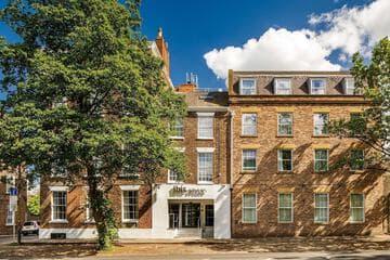 Brick hotel with large windows and white entrance, flanked by bare trees, under a blue sky with scattered clouds.