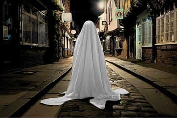 Cobbled street at night with a ghostly figure draped in white cloth, surrounded by historic buildings.