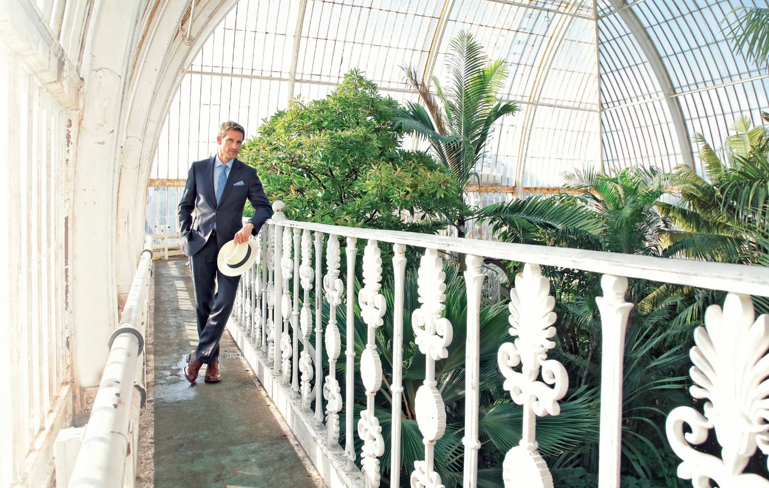 Man in a suit holding a hat, walking on a balcony inside a glass conservatory with lush green plants.
