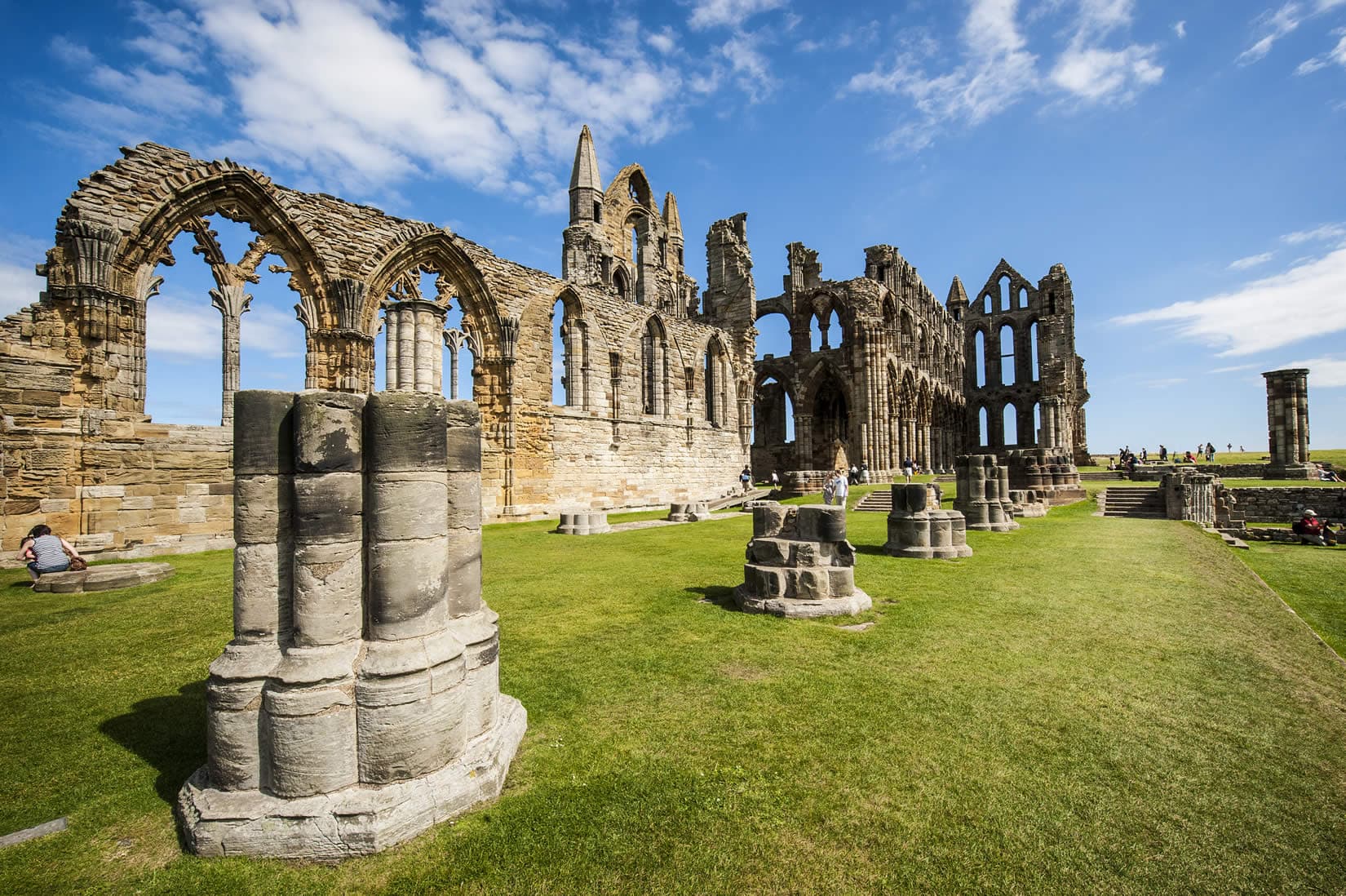 Ruins of Whitby Abbey stand against a dramatic sky, surrounded by grassy fields and a calm water reflection.