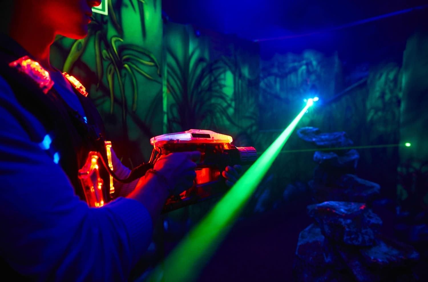 Person playing laser tag in a dark, neon-lit arena with vibrant green and blue lights and jungle-themed walls.