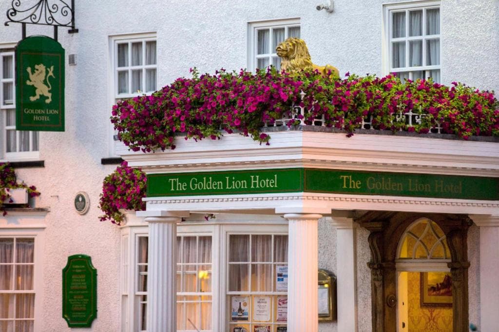 White hotel facade with floral balcony, green signage, golden lion statue, and arched entrance with columns.