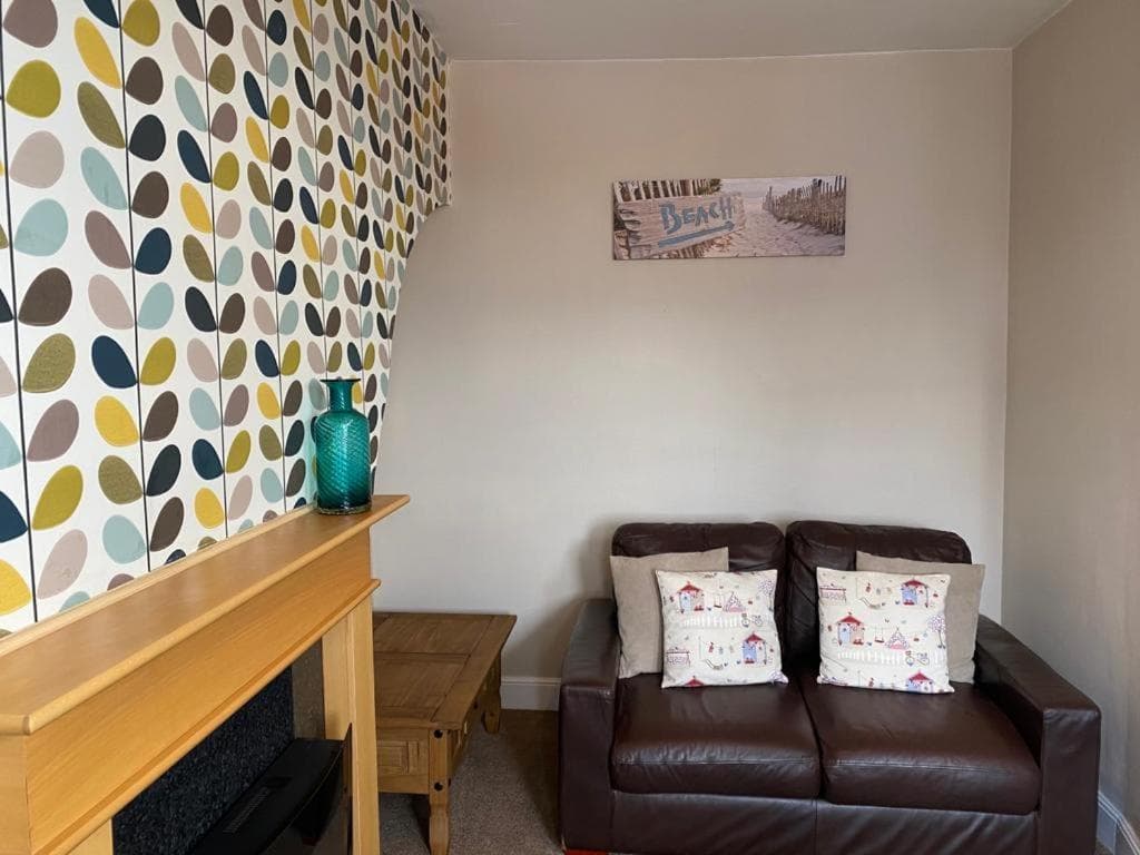 Cozy living room with patterned leaf wallpaper, brown leather sofa, wooden fireplace, and a teal vase on a wooden table.