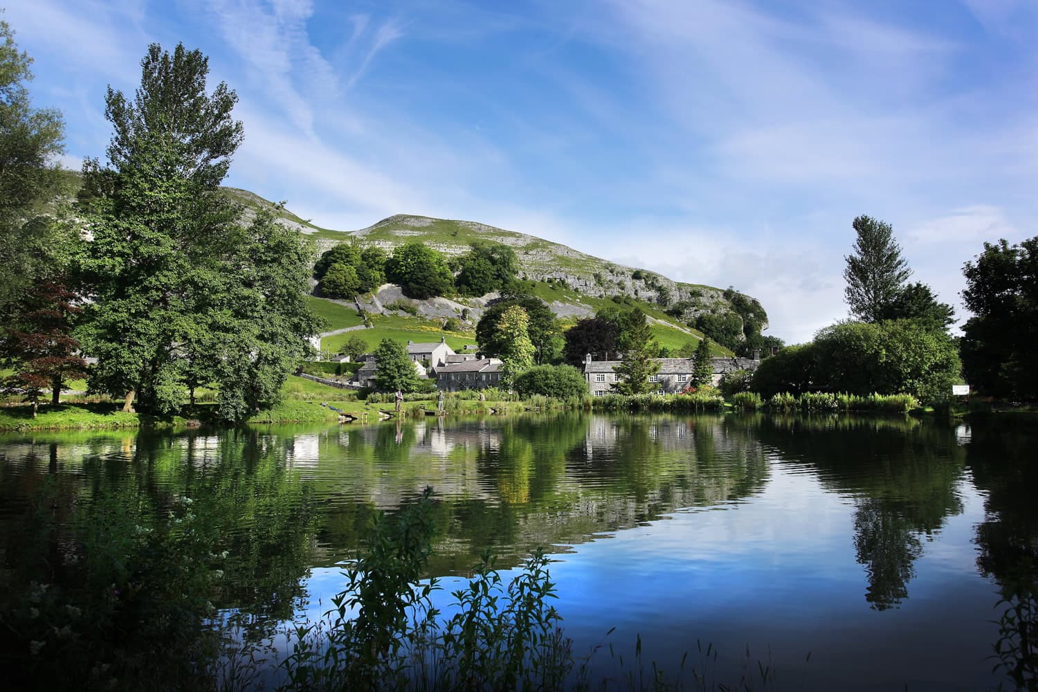 Tranquil lake reflecting lush trees and a hillside, with stone cottages nestled in the landscape under a blue sky.