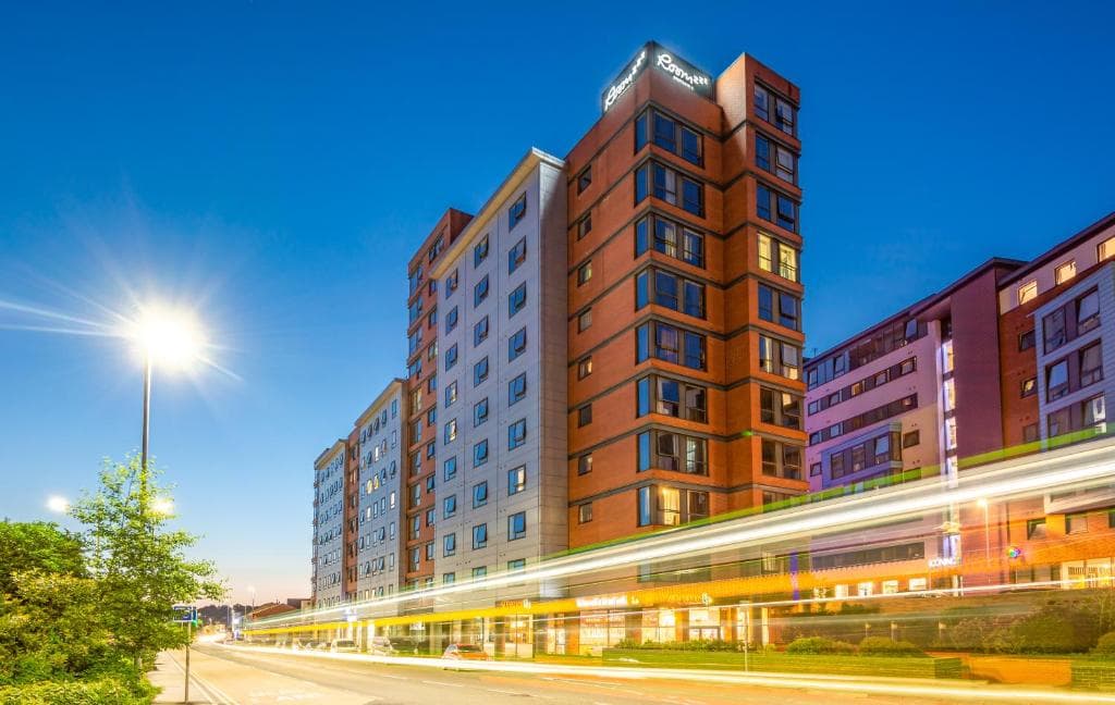 Roomzzz Leeds City West at dusk, tall modern building with bright lights and a streak of passing car lights.