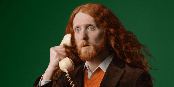 A surprised man with long red hair and a beard holds a vintage telephone, set against a vibrant green background.