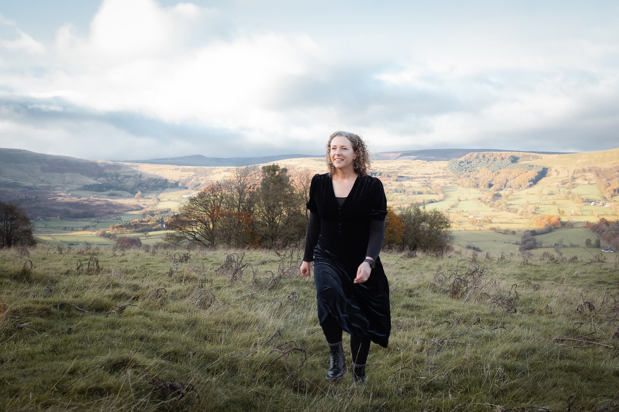 A smiling woman in a black dress walks through green hills, with rolling landscapes and a cloudy sky of North Derbyshire.