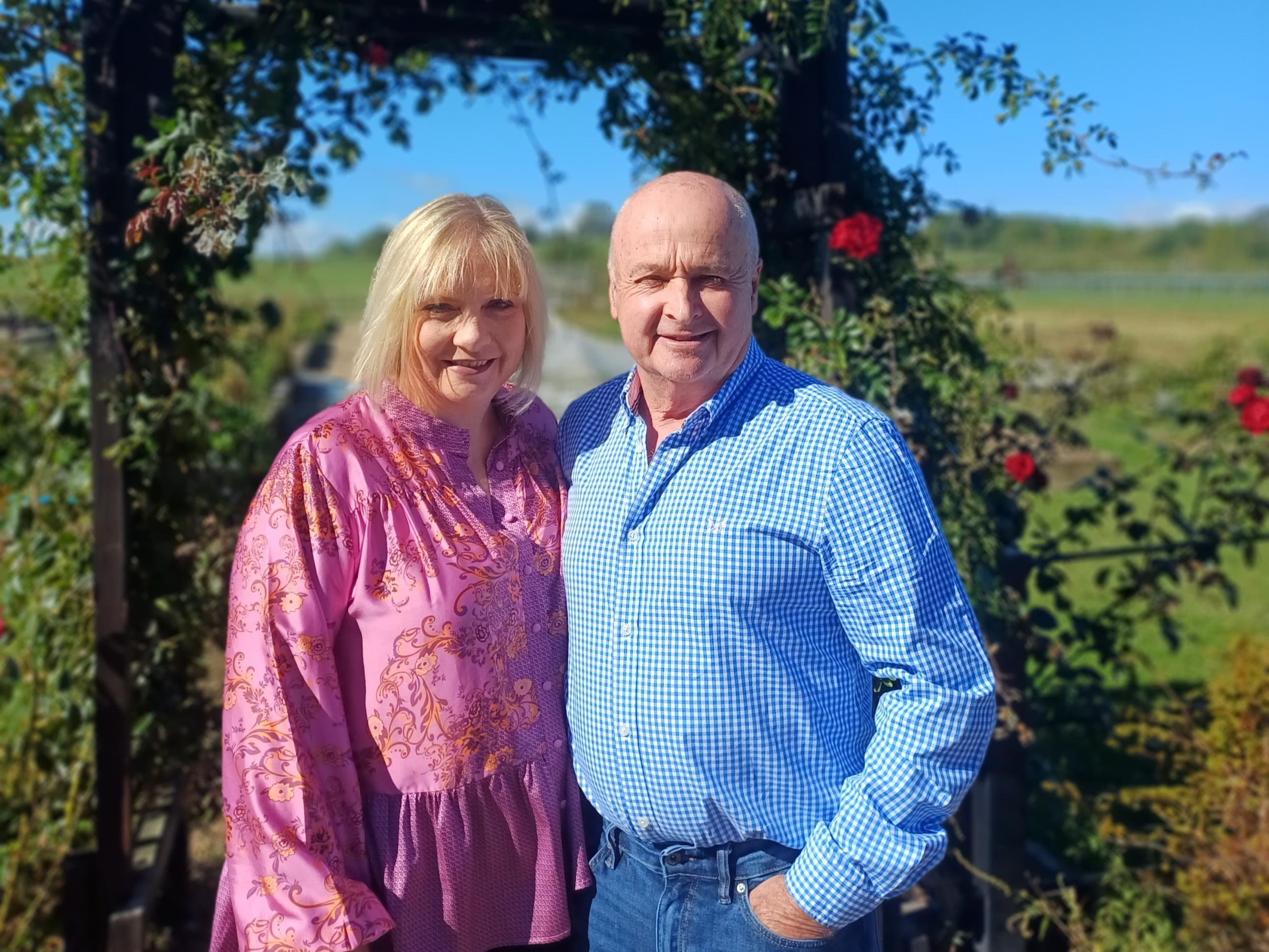 A couple stands together in a sunny garden at Cedarbarn, surrounded by blooming roses and lush green fields.