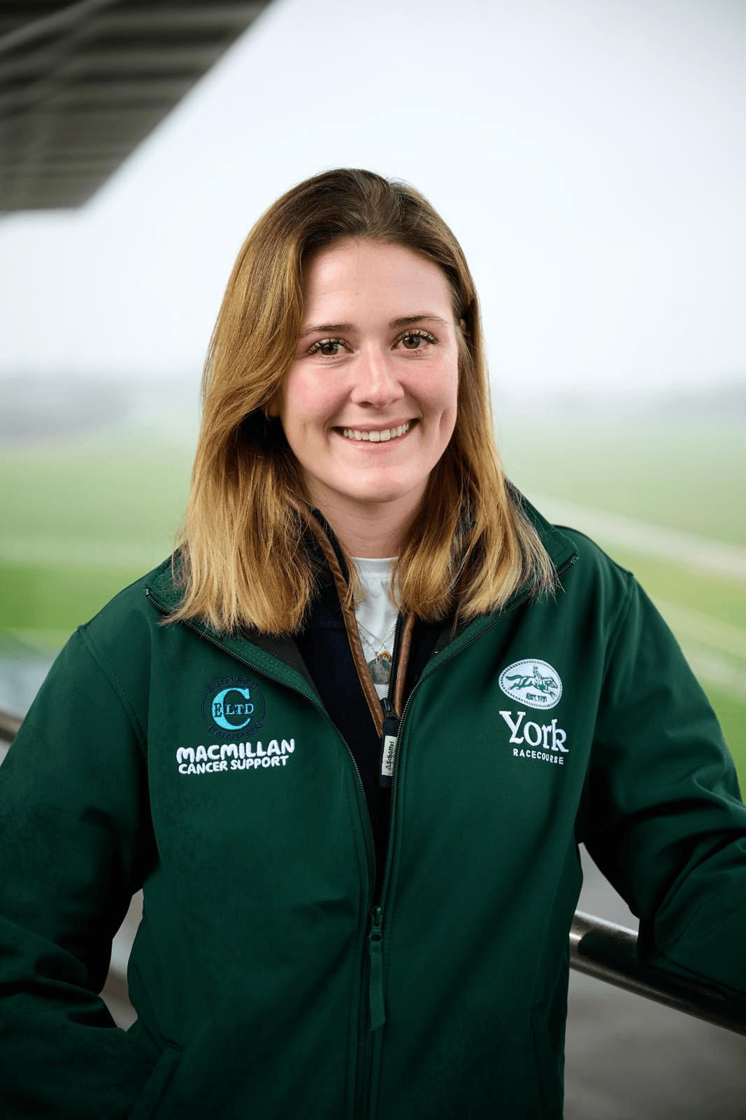 Smiling woman in a green jacket, standing at a racecourse with a foggy landscape in the background, promoting Macmillan Cancer Support.