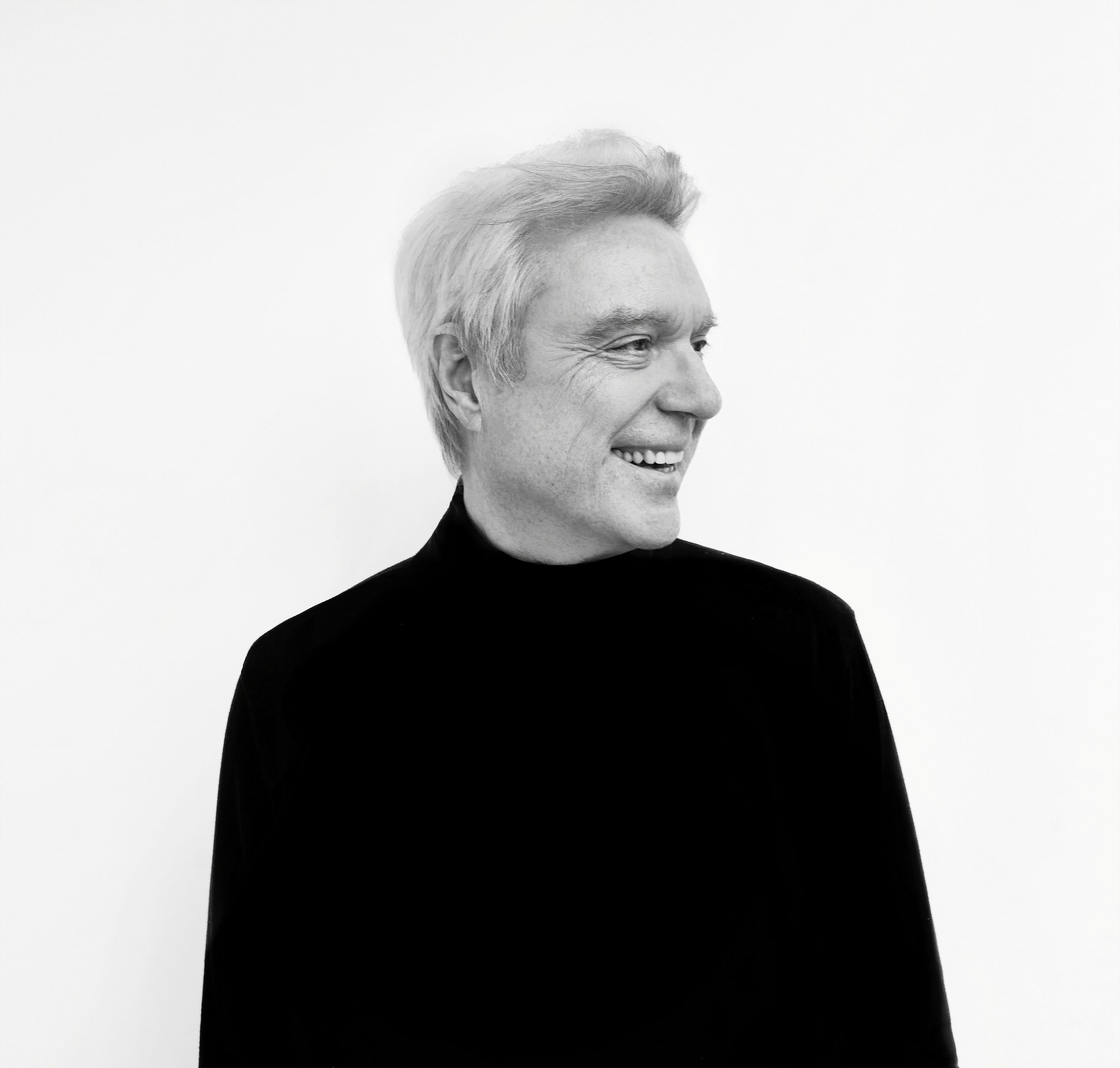 David Byrne smiling in a black turtleneck, exuding a joyful atmosphere, set against a simple white background.