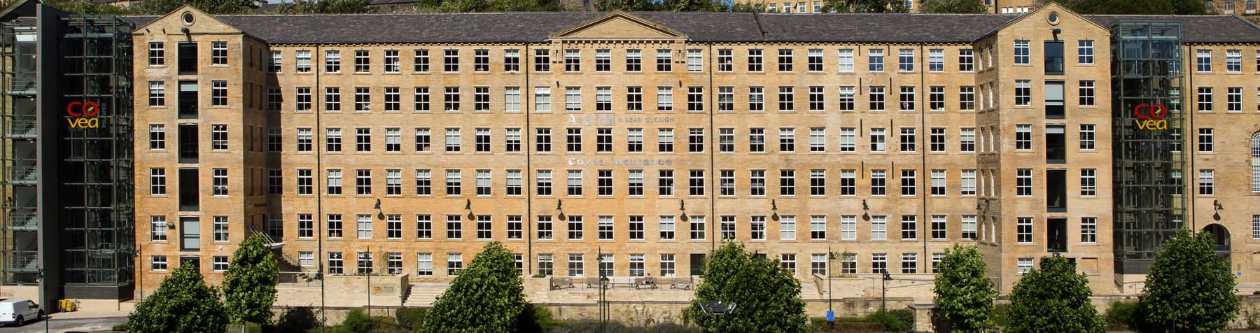 Historic Dean Clough mill with large windows, modern glass extensions, and lush greenery in front, showcasing urban regeneration.