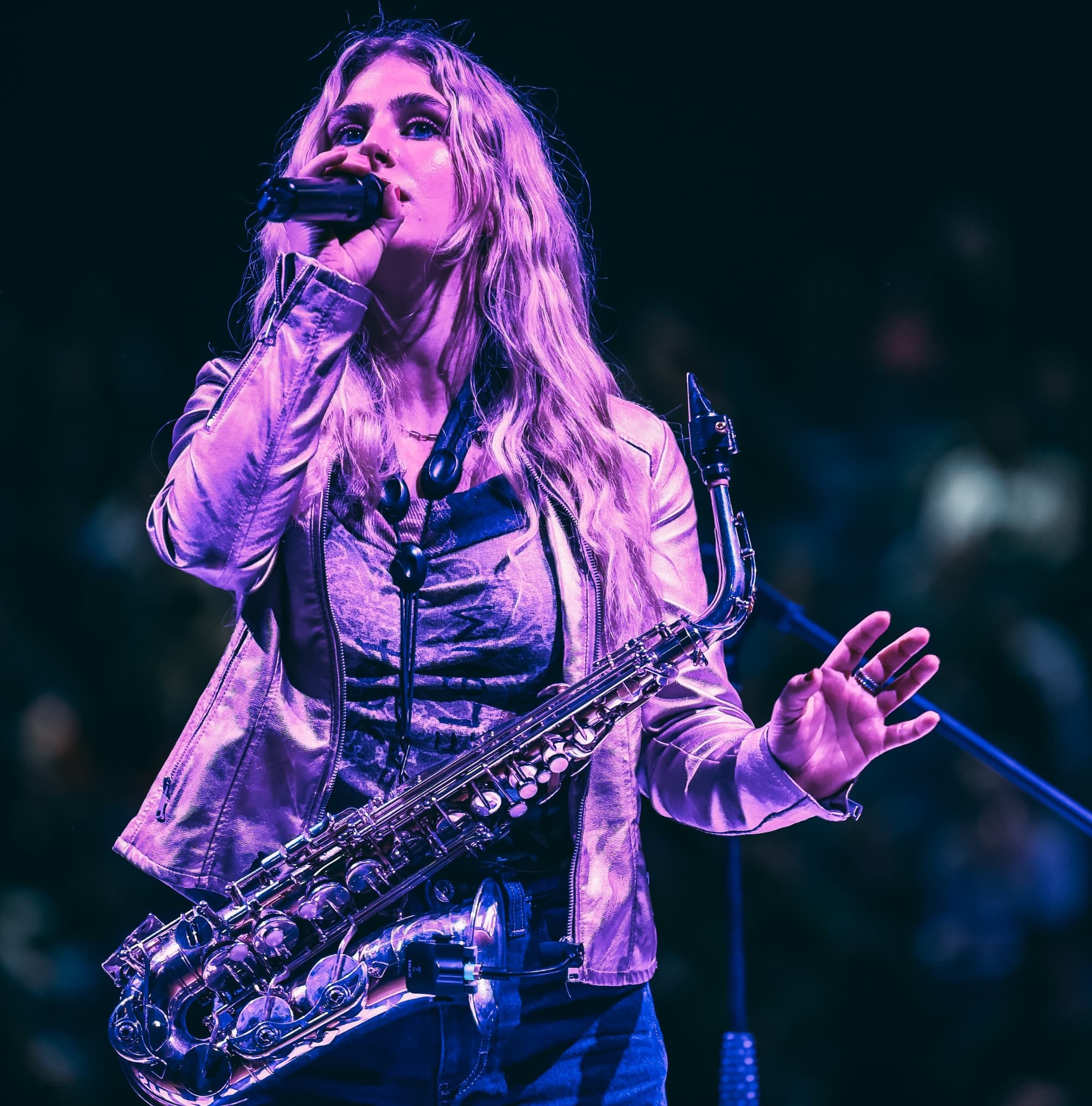 A singer with long hair passionately performs on stage, holding a saxophone, surrounded by an enthusiastic audience.