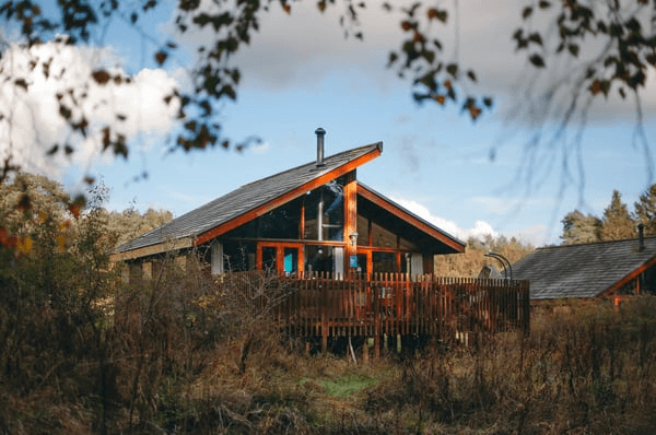 Luxury cabin nestled in lush Yorkshire forest, featuring large windows, a wooden deck, and surrounded by greenery.