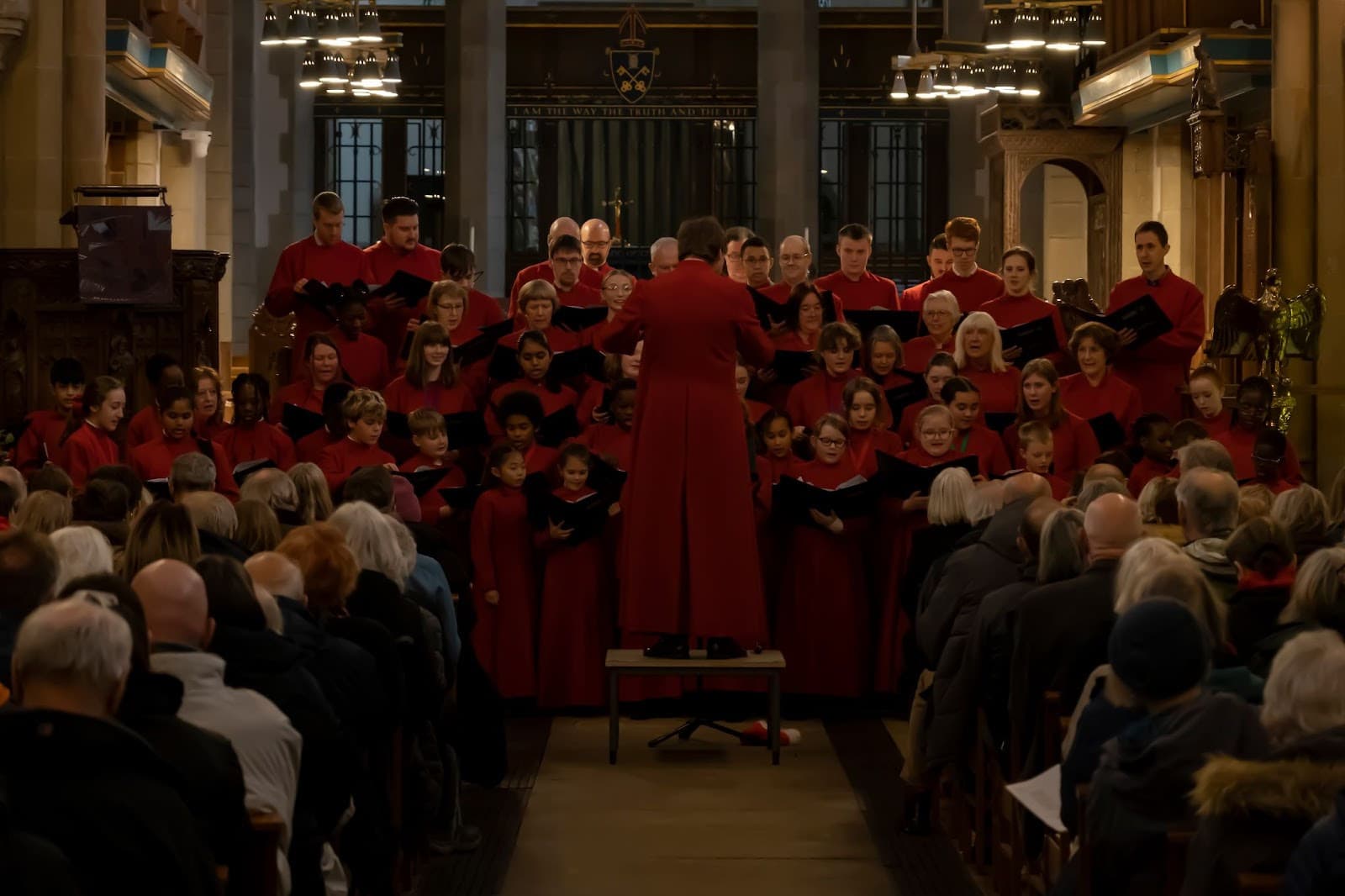 A choir in red robes performs in Bradford Cathedral, surrounded by an attentive audience in a candlelit atmosphere.
