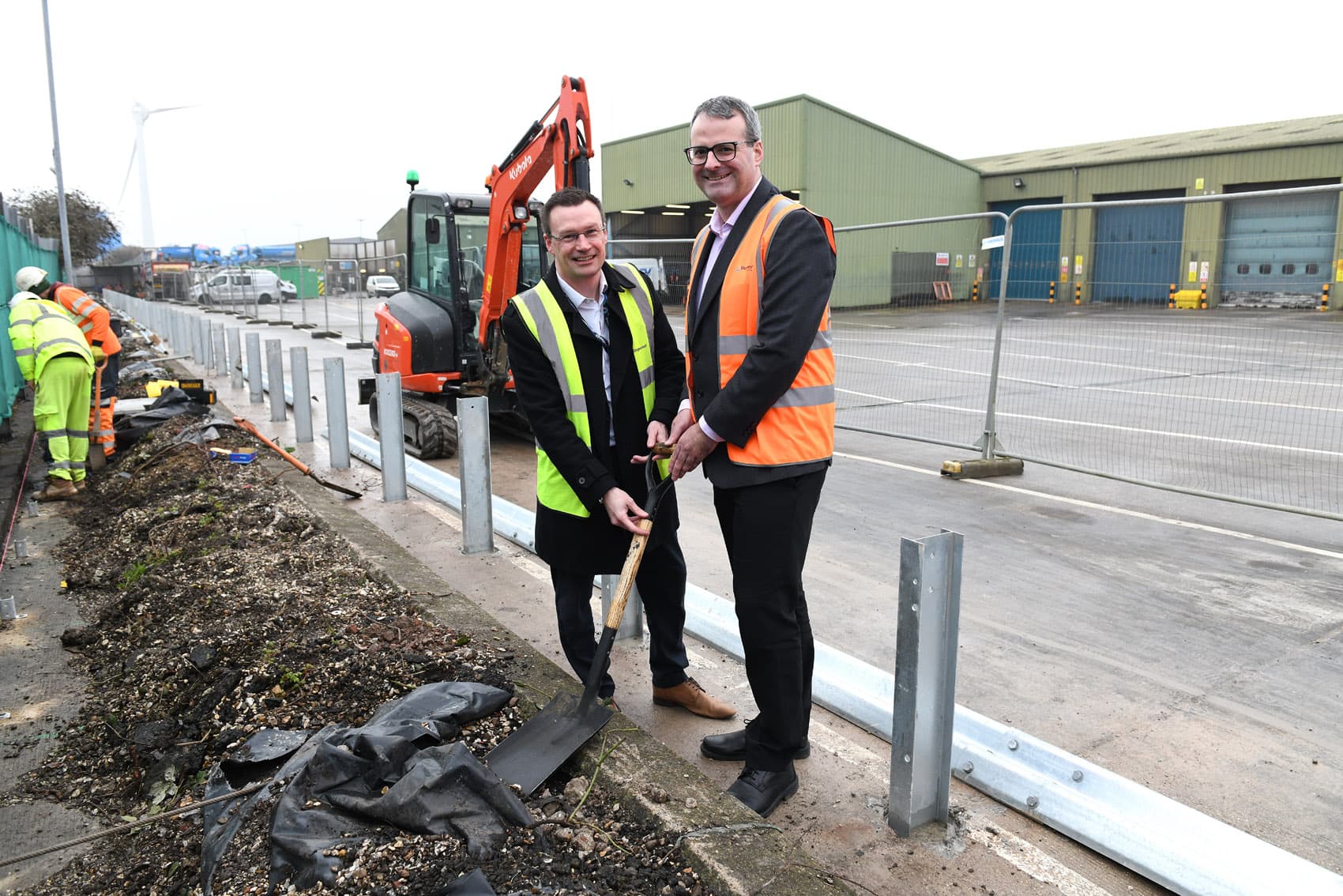 Two men in high-visibility vests break ground at a construction site in Hull, surrounded by workers and machinery.