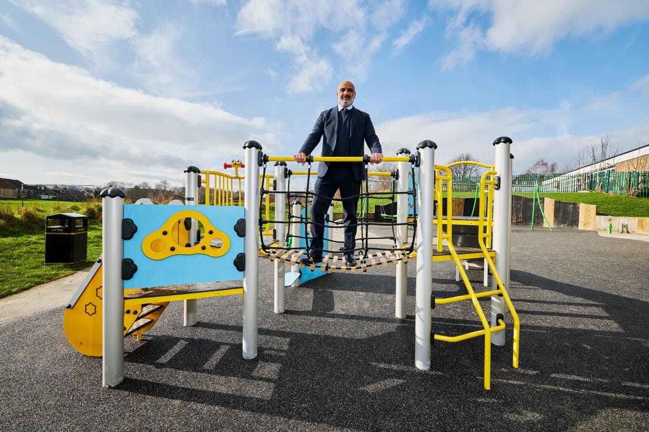 A man in a suit stands on a colorful climbing frame in Kettlethorpe's newly refurbished play area under a bright sky.