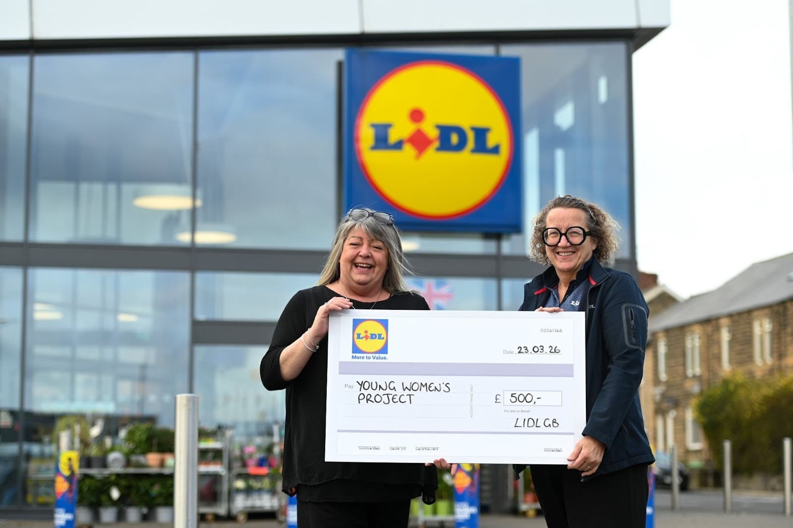 Two women outside a Lidl store in North East England hold a large donation check for a local charity, smiling warmly.