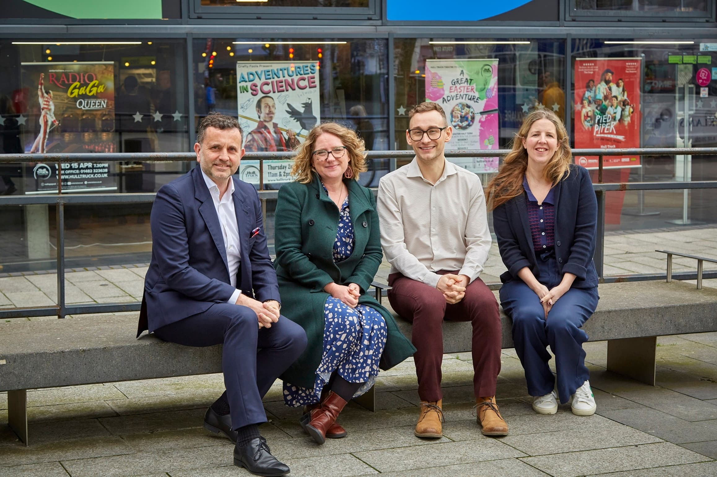 Four professionals sit on a bench outside a vibrant arts venue, with colorful posters in the background showcasing local events.