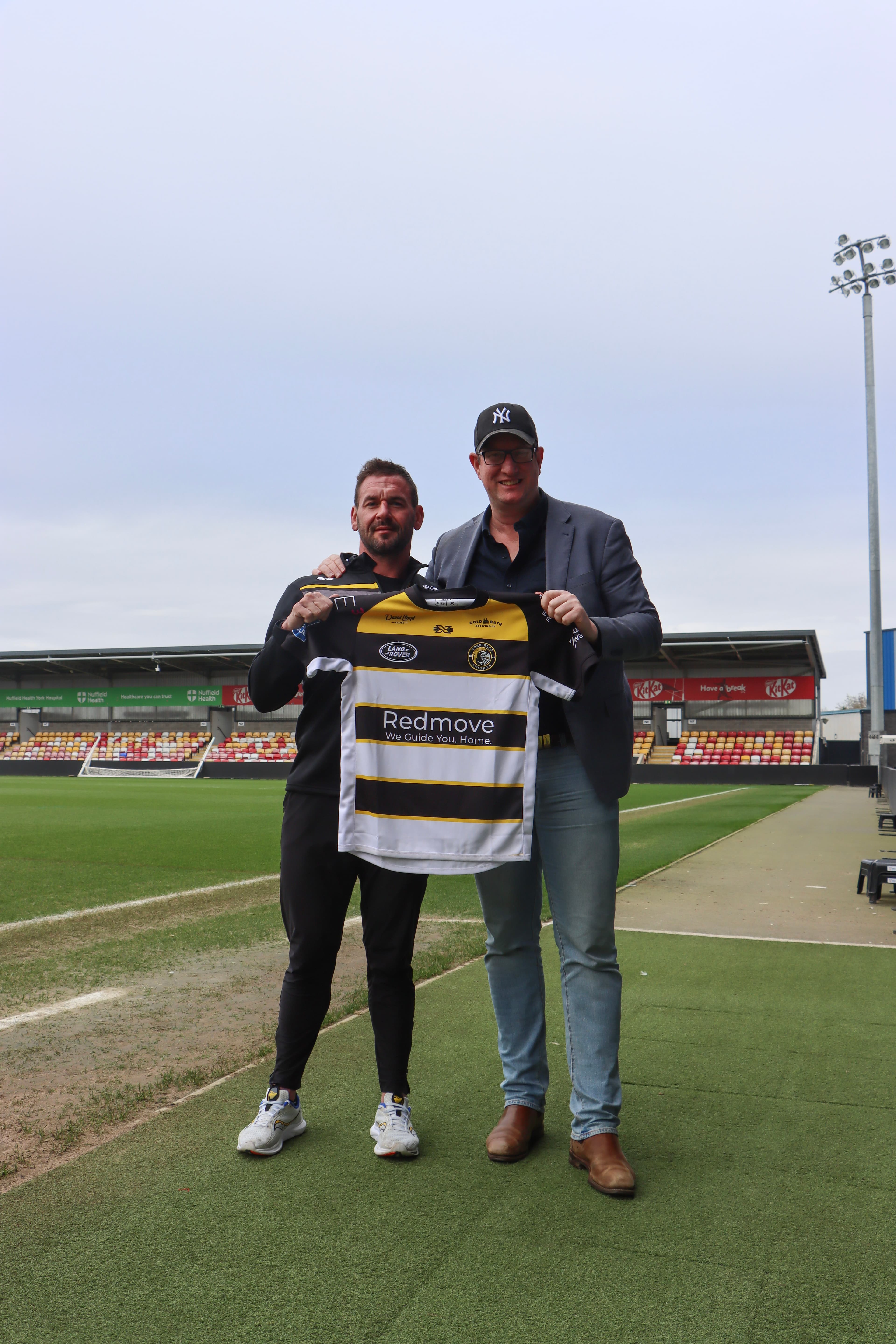 Two men stand on a rugby field, holding a team jersey, with stadium seating and overcast skies in the background.