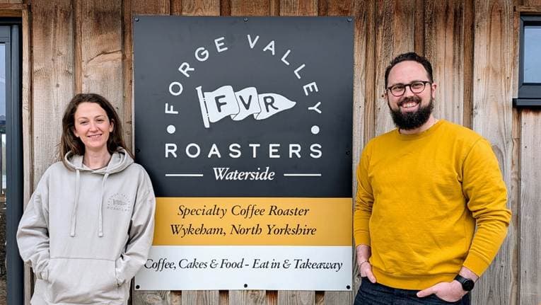Two smiling individuals stand outside the Forge Valley Roasters sign at North Yorkshire Water Park, surrounded by rustic wooden architecture.