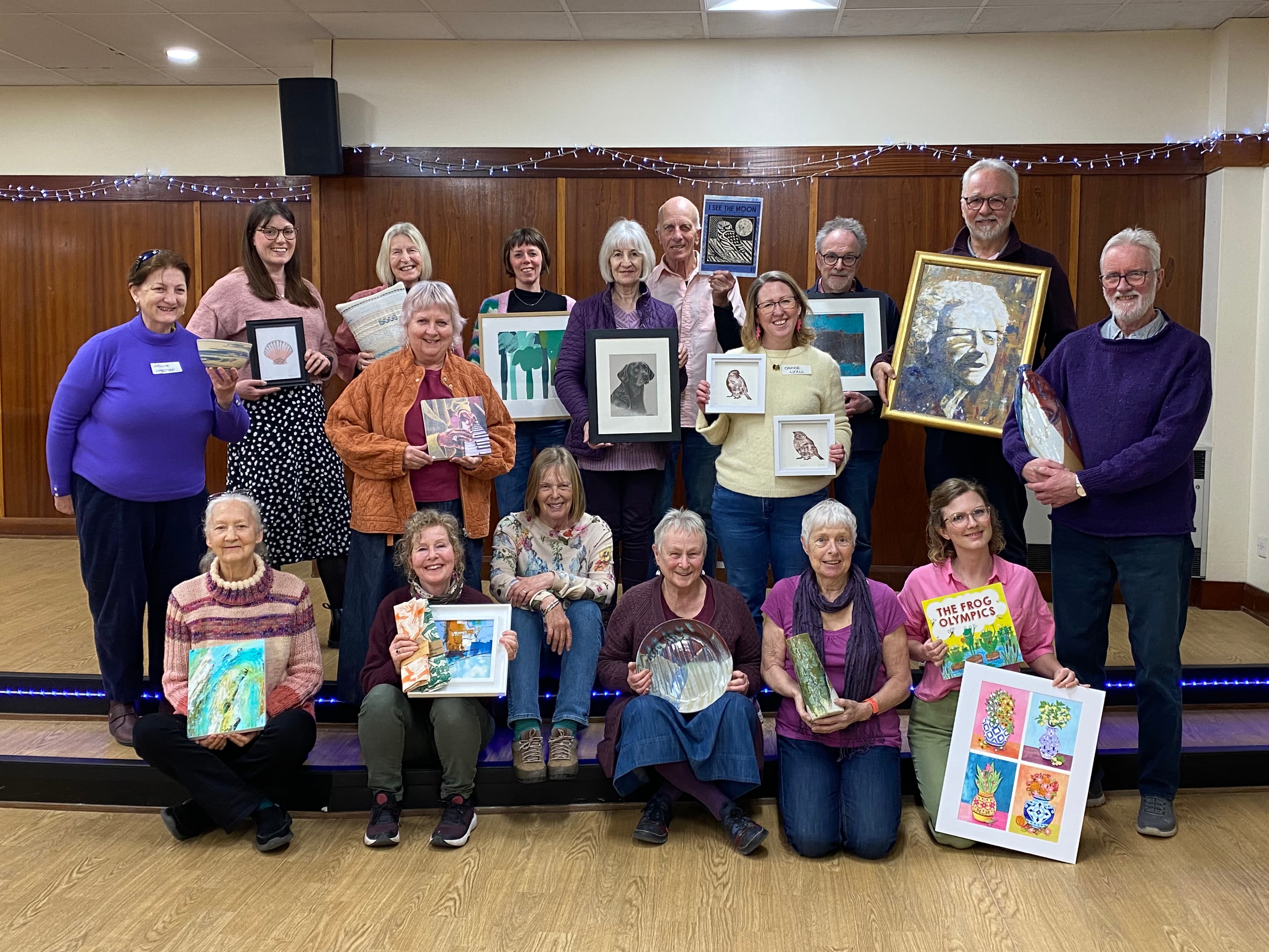 A group of smiling artists in a community hall, holding various artworks, showcasing creativity and collaboration in Pocklington.