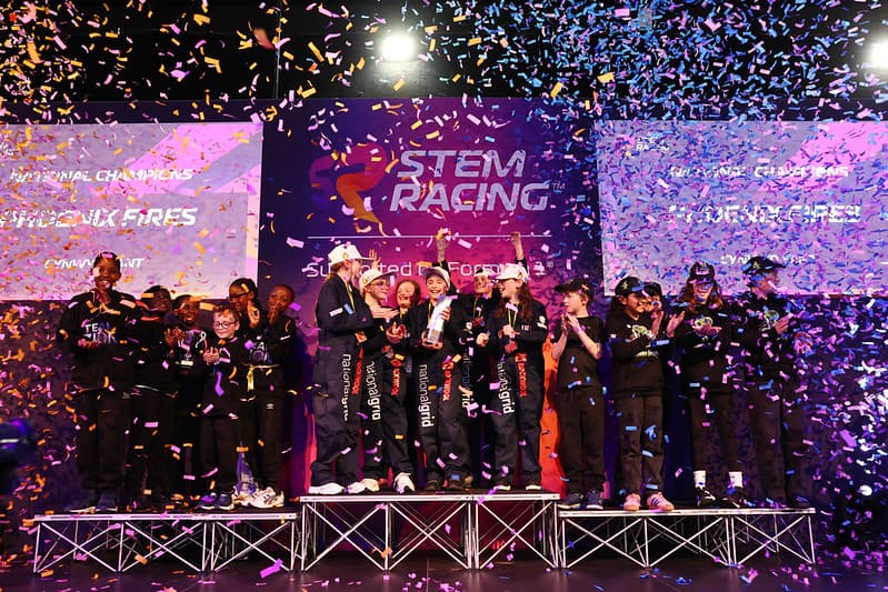 A group of jubilant students celebrate on stage with trophies, surrounded by colorful confetti at the STEM Racing UK Finals.