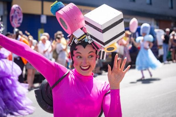 A performer in a vibrant pink costume waves, showcasing a whimsical hat adorned with colorful props at the Pontefract Liquorice Festival.