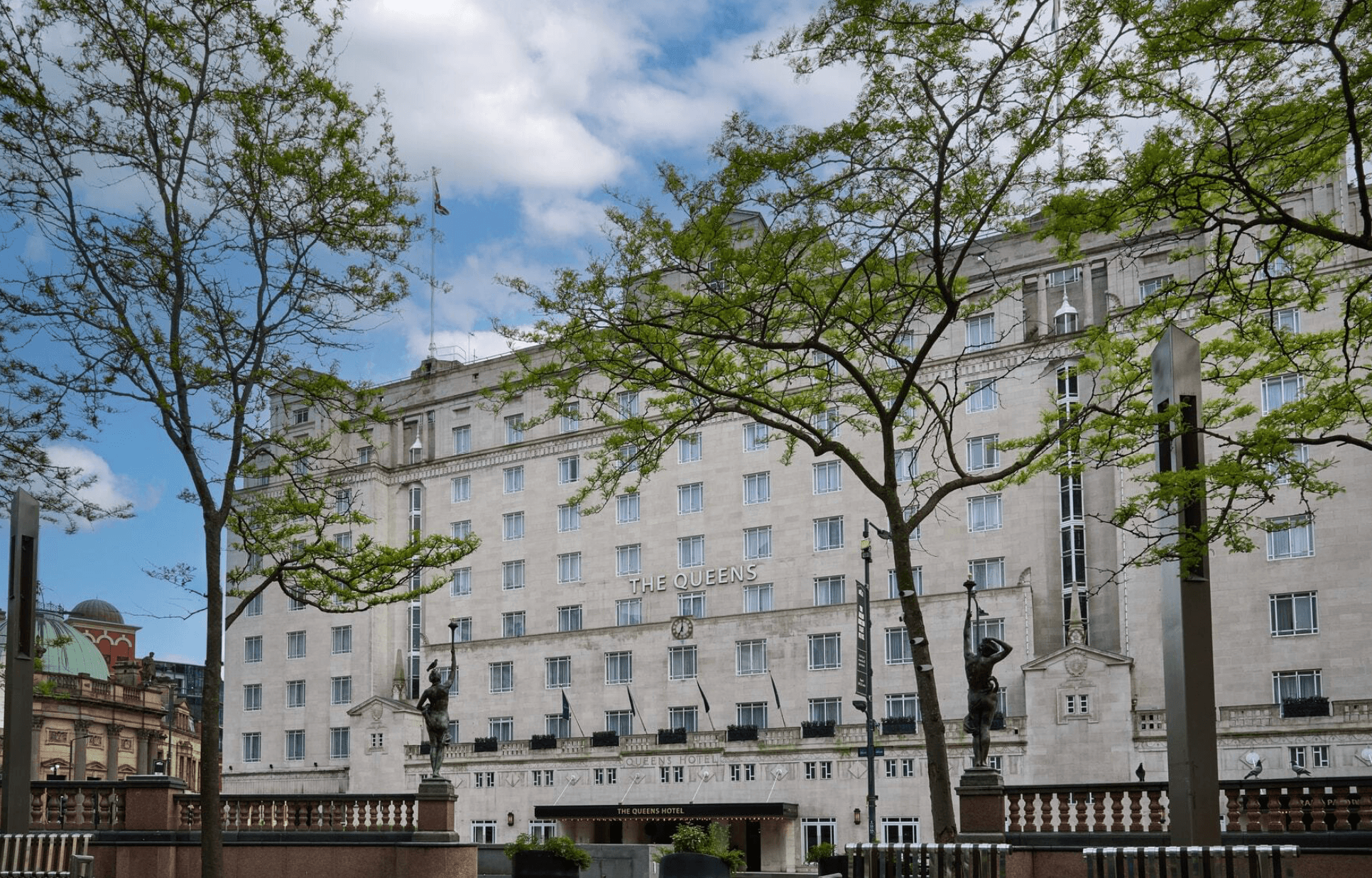 The Queens Leeds hotel stands majestically with greenery in the foreground and a bright blue sky above.