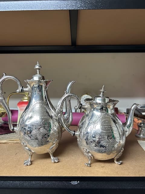 Two ornate silver jugs with intricate engravings, resting on a wooden surface, reflecting light and showcasing craftsmanship.