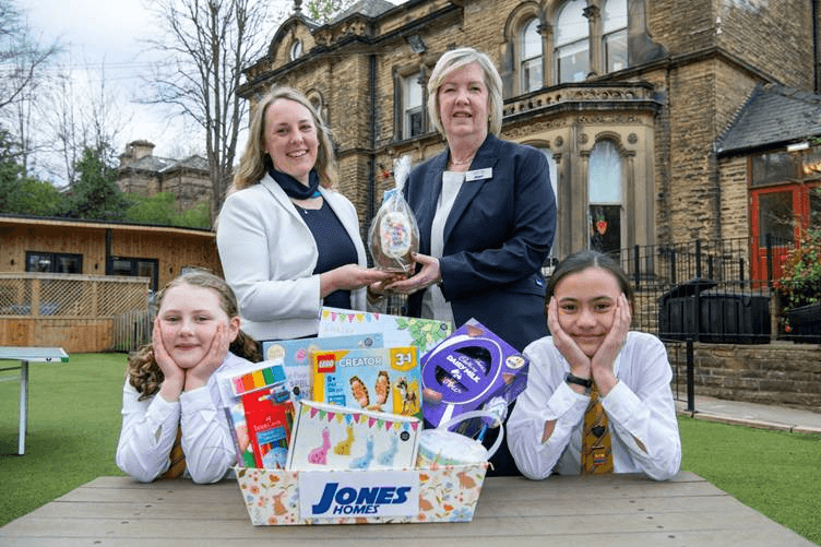 Two women hold an Easter egg, flanked by two schoolgirls, with a historic stone building and green lawn in the background.