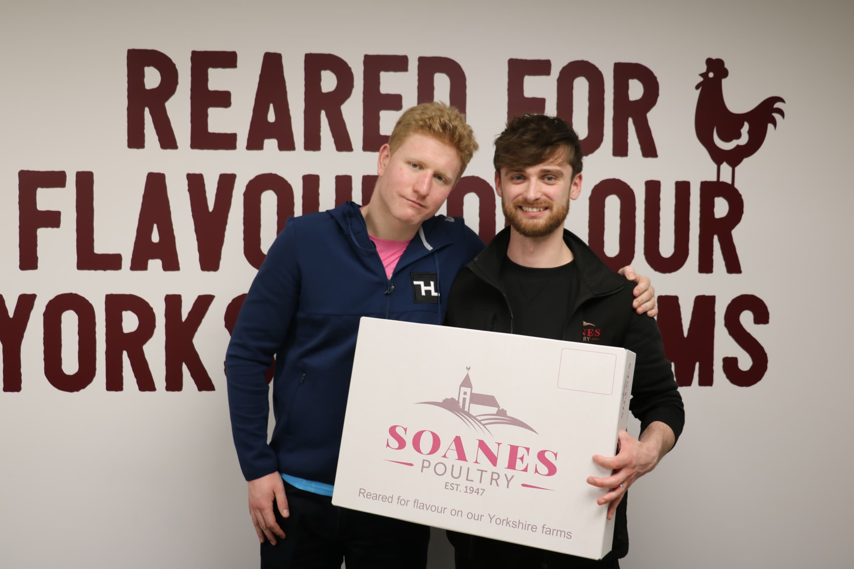 Two young men stand inside a business with a sign reading "Reared for flavour on your Yorkshire farms," holding a Soanes Poultry box.