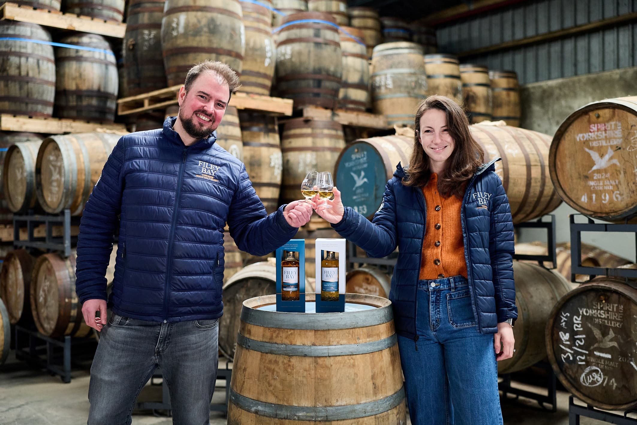 Two distillers toast with glasses of whisky in a rustic distillery filled with wooden barrels and vibrant atmosphere.