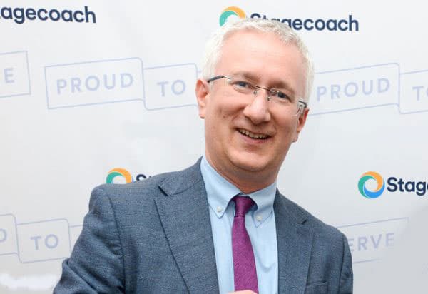 A smiling man in a suit stands in front of a backdrop featuring Stagecoach and Global's Make Some Noise logos.