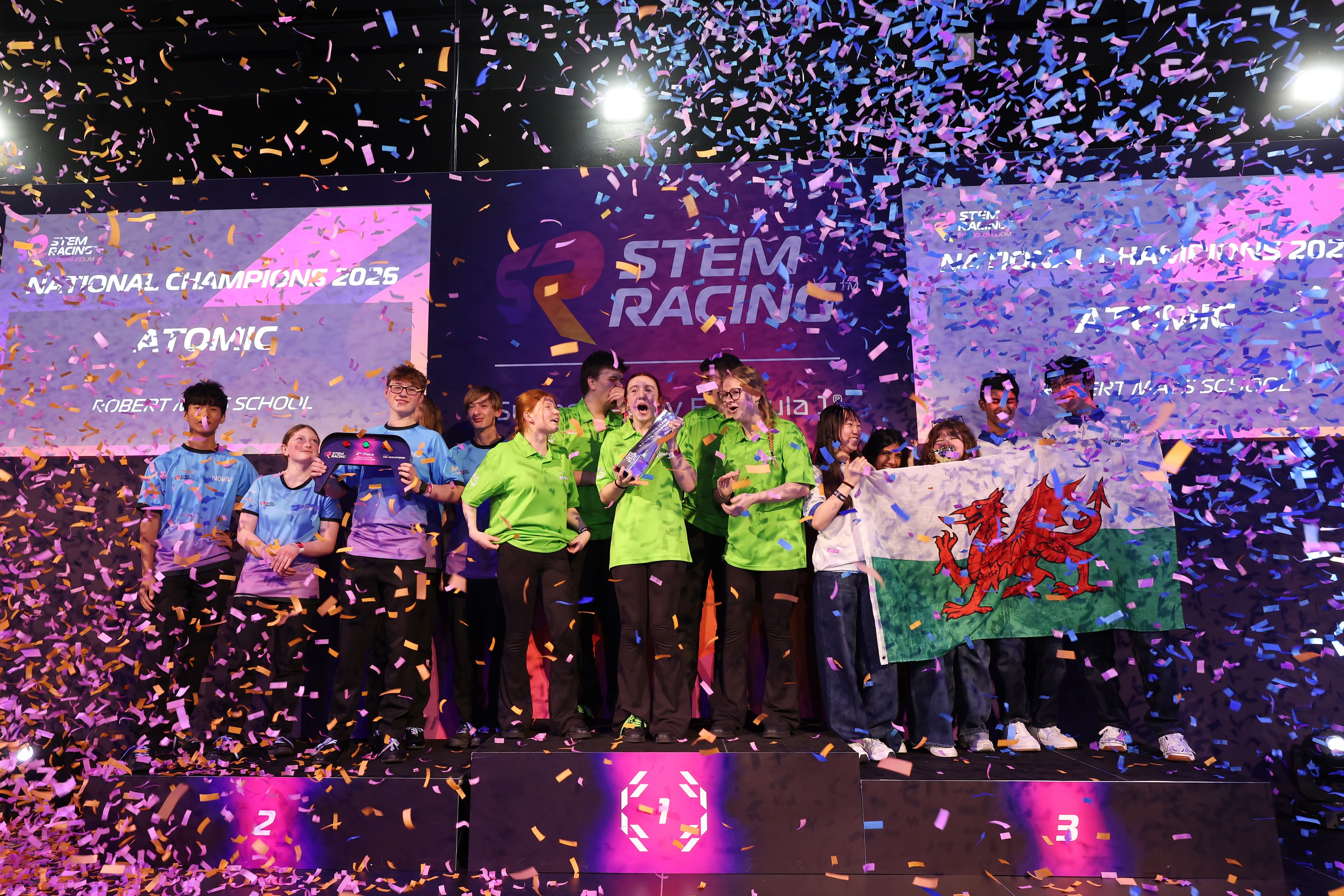 Celebratory scene with confetti as students in colorful shirts hold a trophy and a Welsh flag on stage at a STEM event.