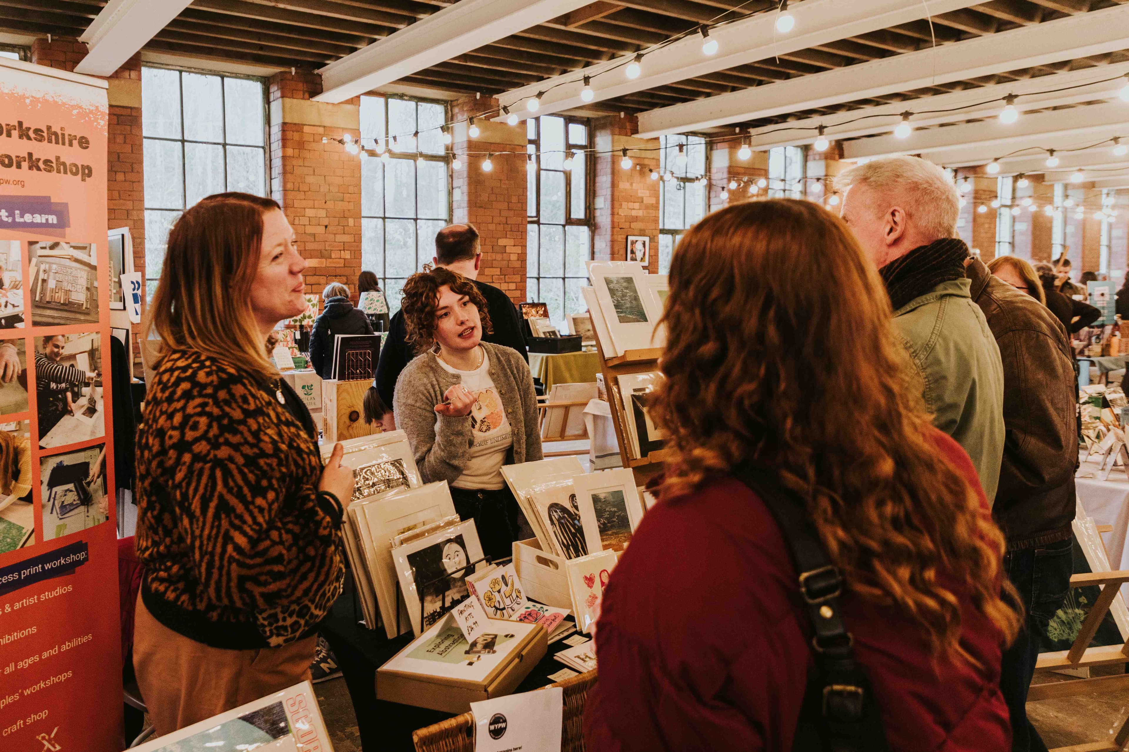 A vibrant print fair at Sunny Bank Mills, with attendees engaging around colorful stalls and artwork in a historic industrial space.