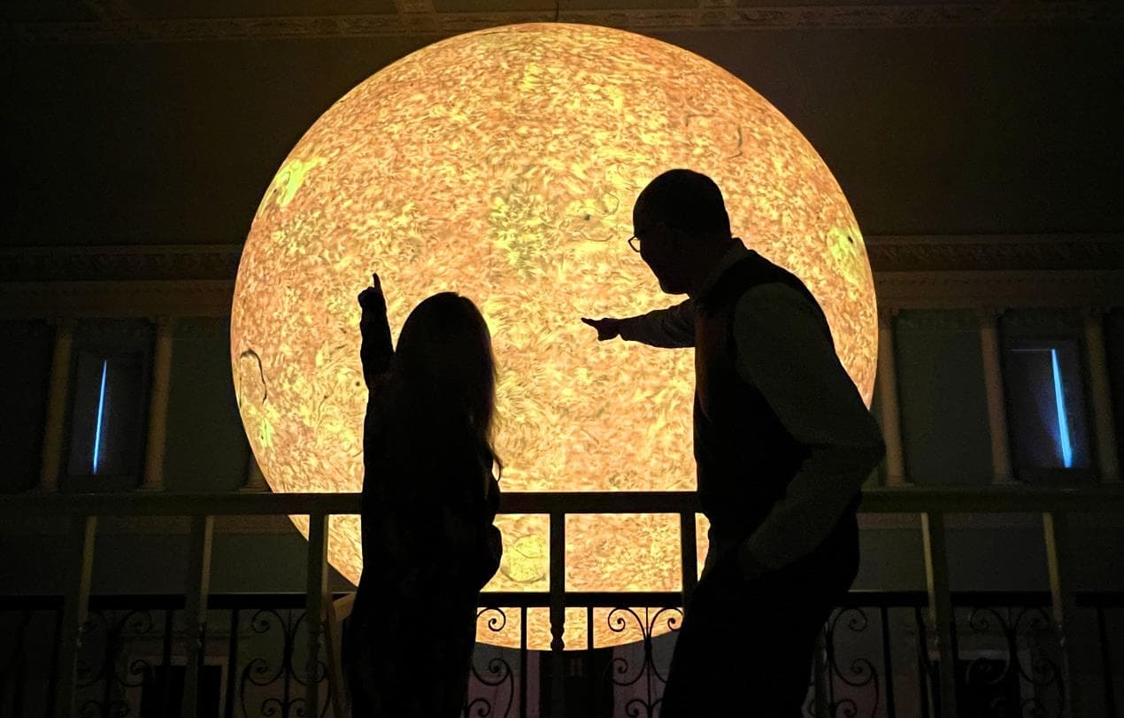 Two visitors point at a large, glowing sun installation in a grand indoor space, creating a warm, inviting atmosphere.