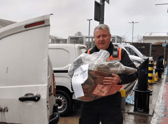 A man in work attire holds a large bag of seized items near a white van in a city setting, with grey skies above.