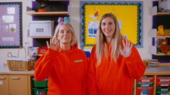 Two smiling women in bright orange hoodies wave from a classroom, surrounded by colorful educational materials.