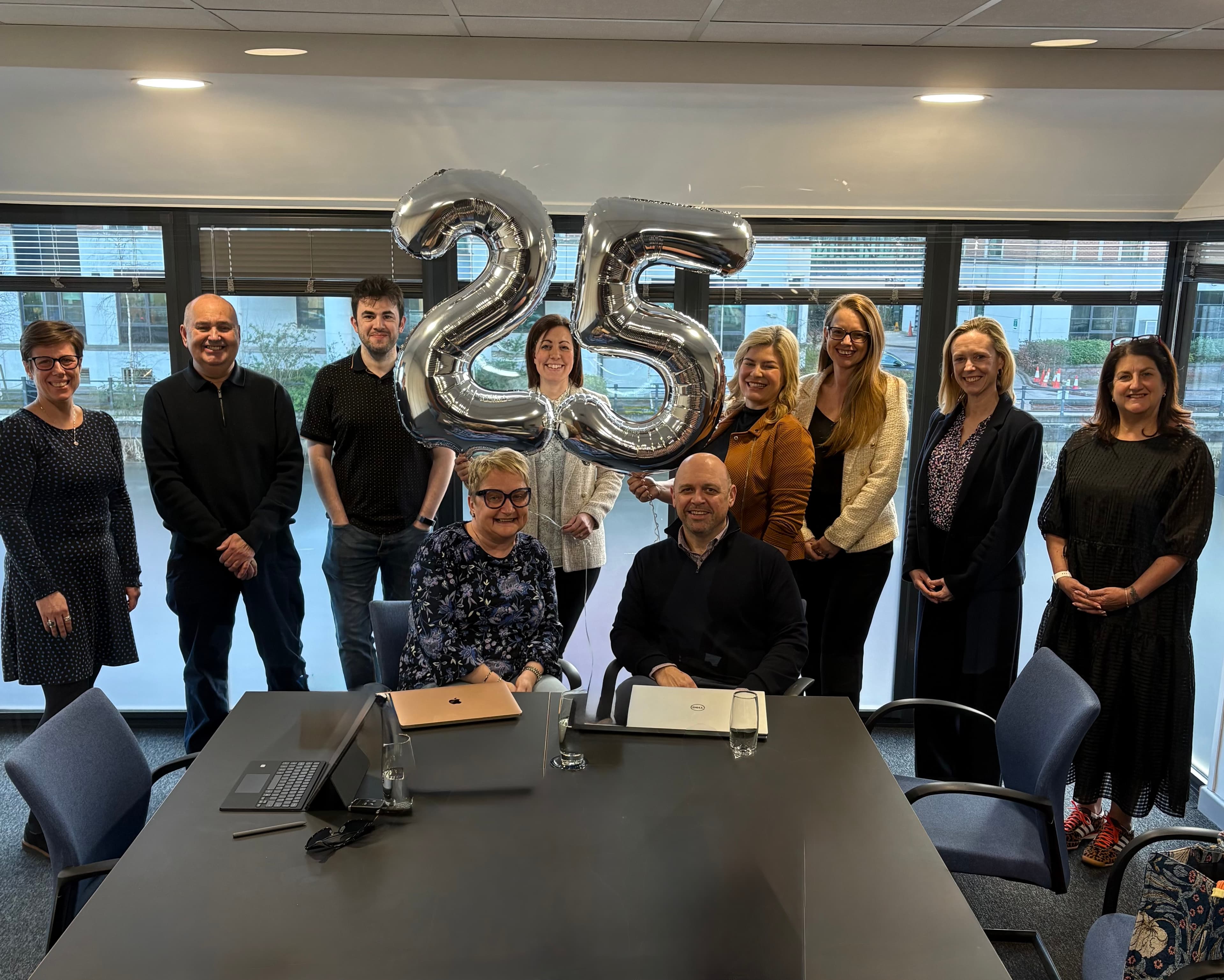 A group of professionals in a modern office celebrate 25 years of supporting York businesses with large silver balloons.