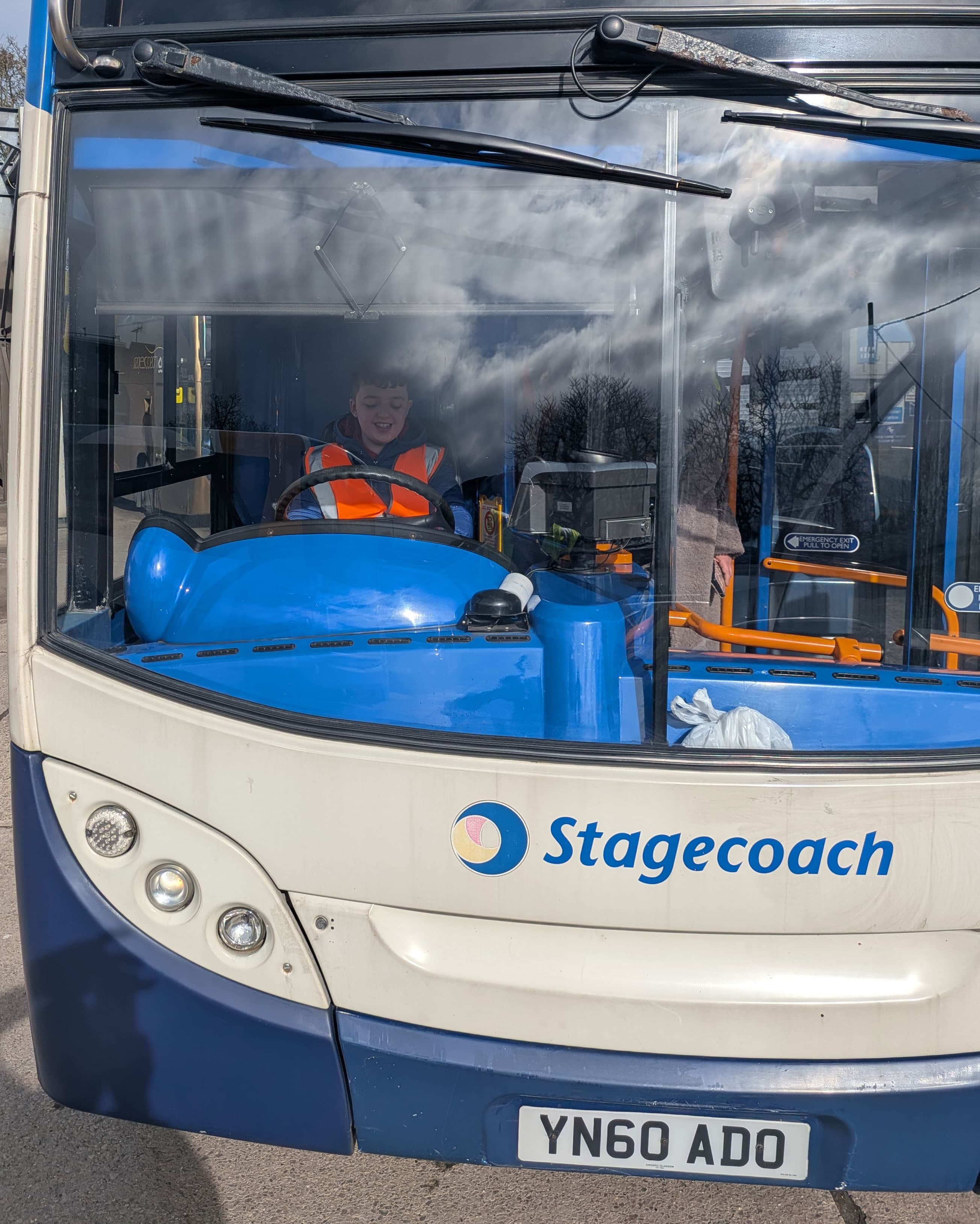 A young boy in a high-visibility vest sits in the driver's seat of a Stagecoach bus at Sheffield depot, smiling.