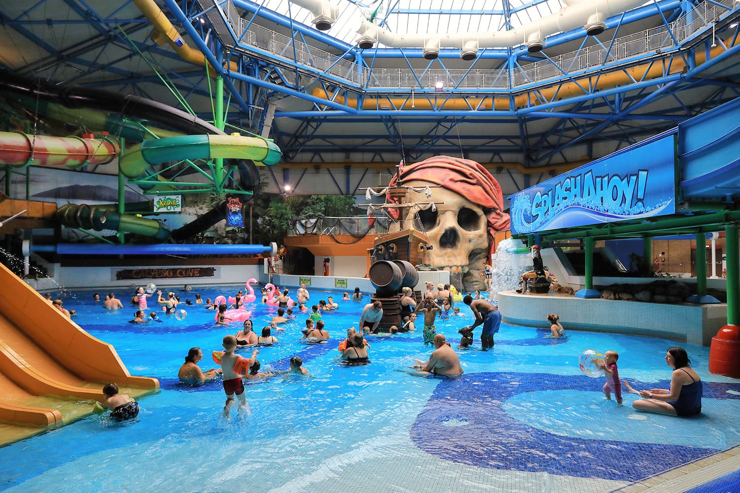 Indoor water park with pirate-themed slides, large skull structure, and families enjoying pools under a glass roof.
