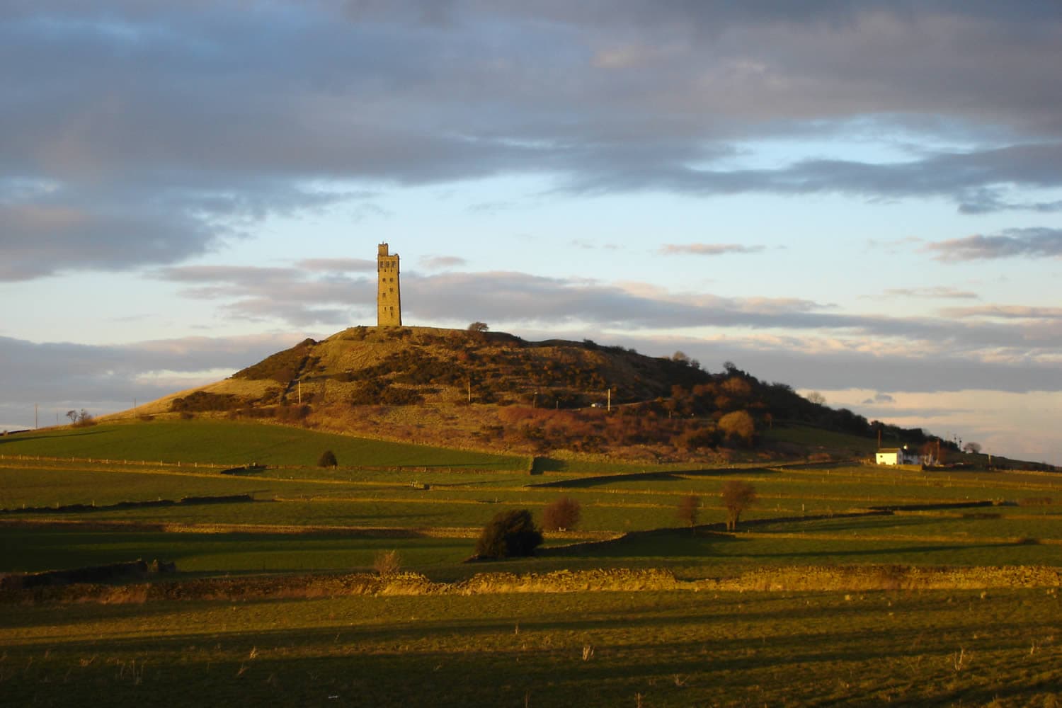Victoria Tower atop Castle Hill, surrounded by rolling green fields under a dramatic cloudy sky.