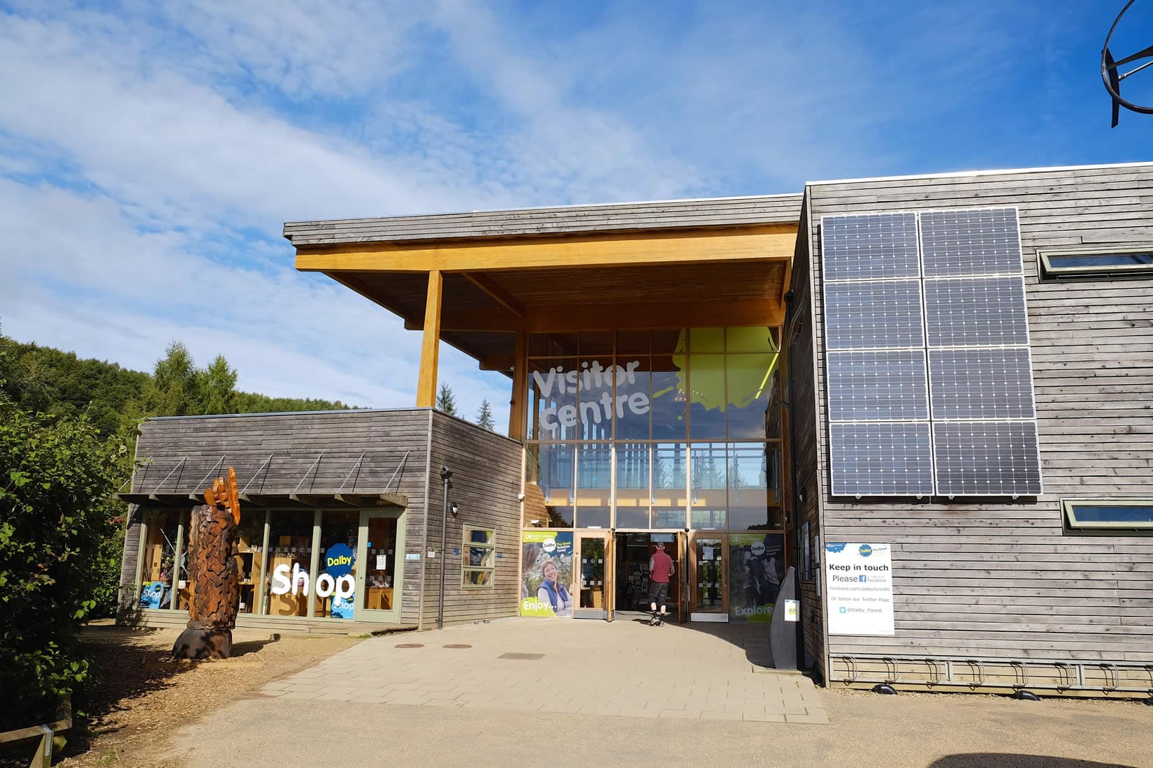 Modern wooden visitor center with large glass windows, solar panels, and people entering under a clear blue sky.