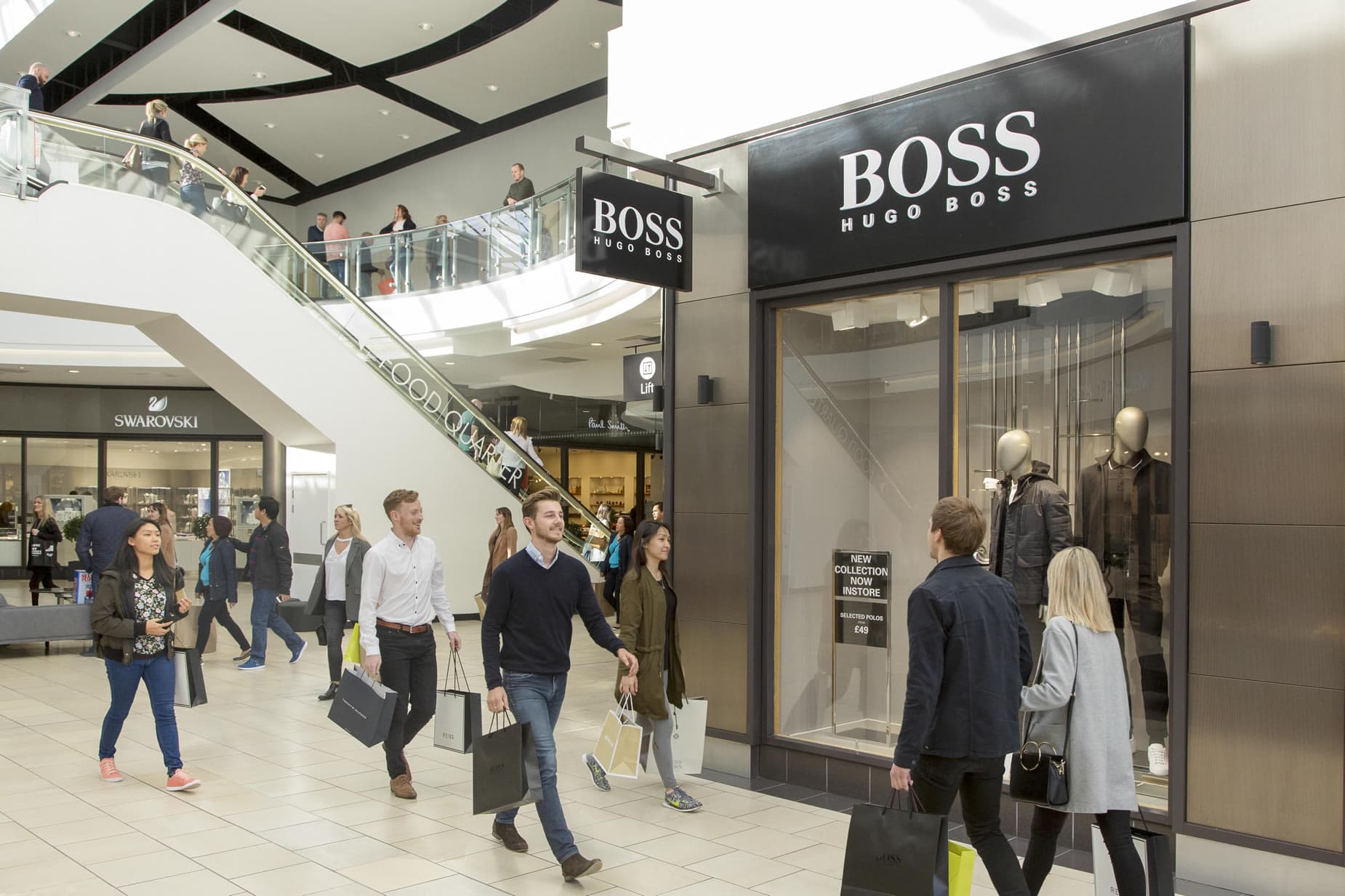 Shoppers with bags walk past Hugo Boss store in a modern, bright shopping mall with escalator and glass railings.
