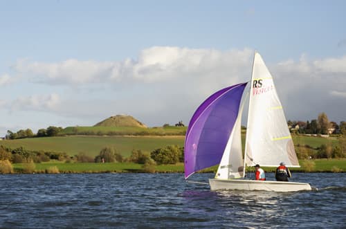 Sailboat on a lake with green hills and a distant mound under a partly cloudy sky at Pugneys Country Park.