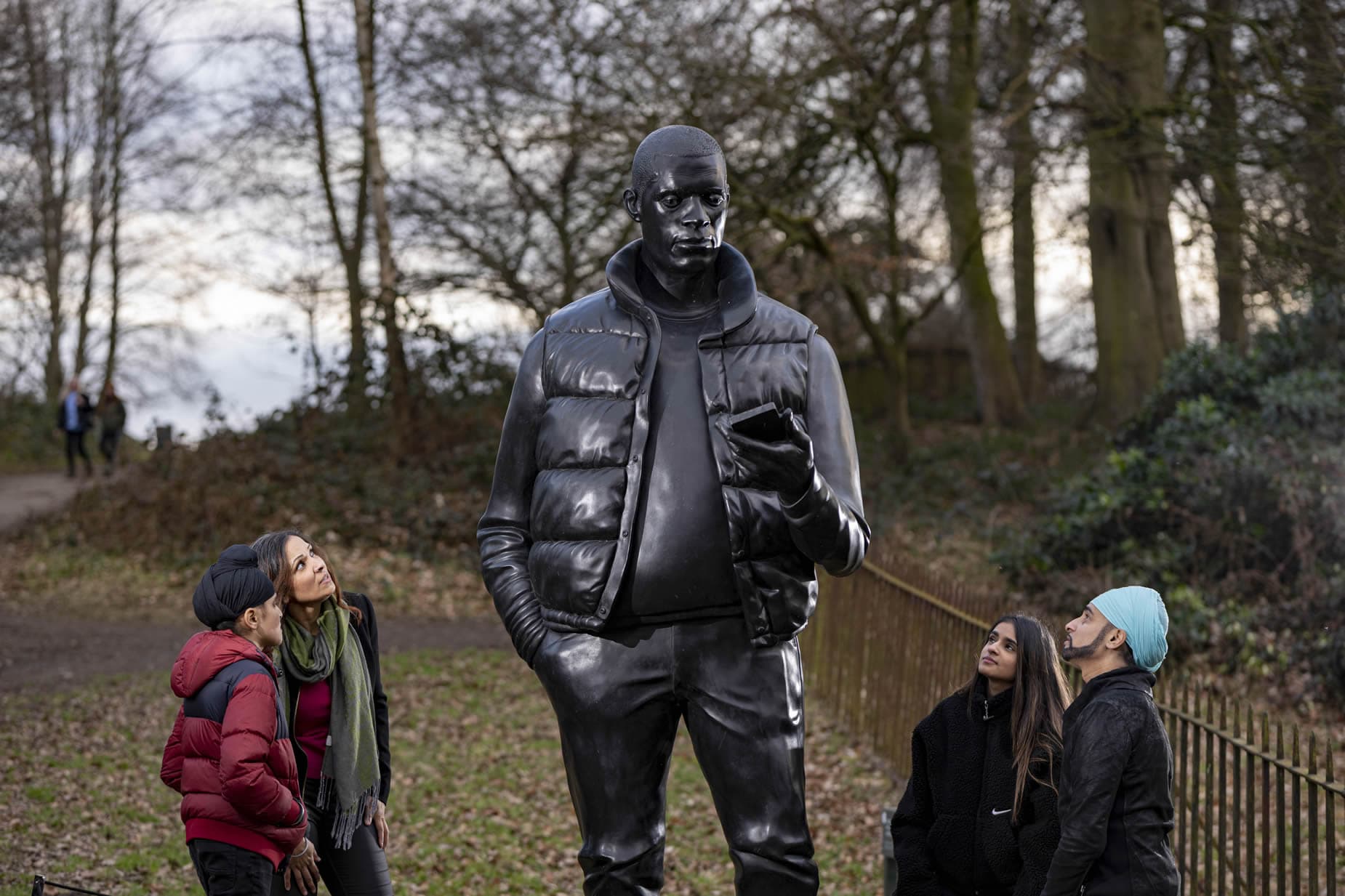 Visitors admire a towering sculpture of a man in a jacket, set in a wooded park landscape with a cloudy sky.