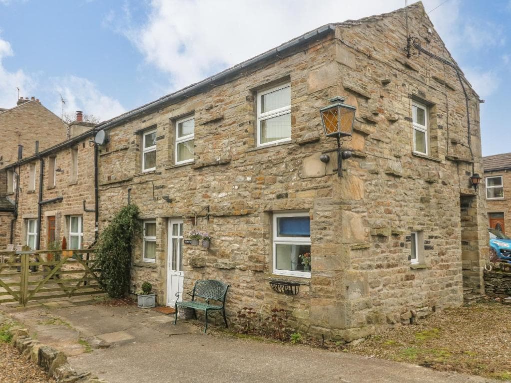 Rustic stone cottage with white-framed windows, wooden door, and wrought iron bench in a quaint village setting.