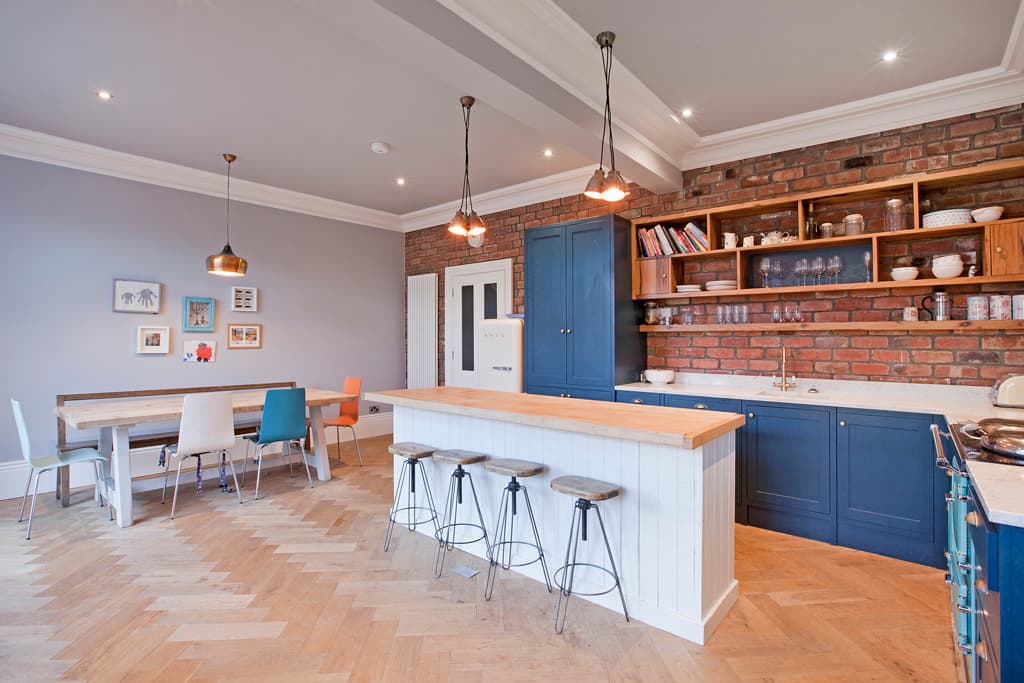 Modern kitchen with blue cabinetry, wooden dining table, colorful chairs, and exposed brick wall. Stylish lighting fixtures.