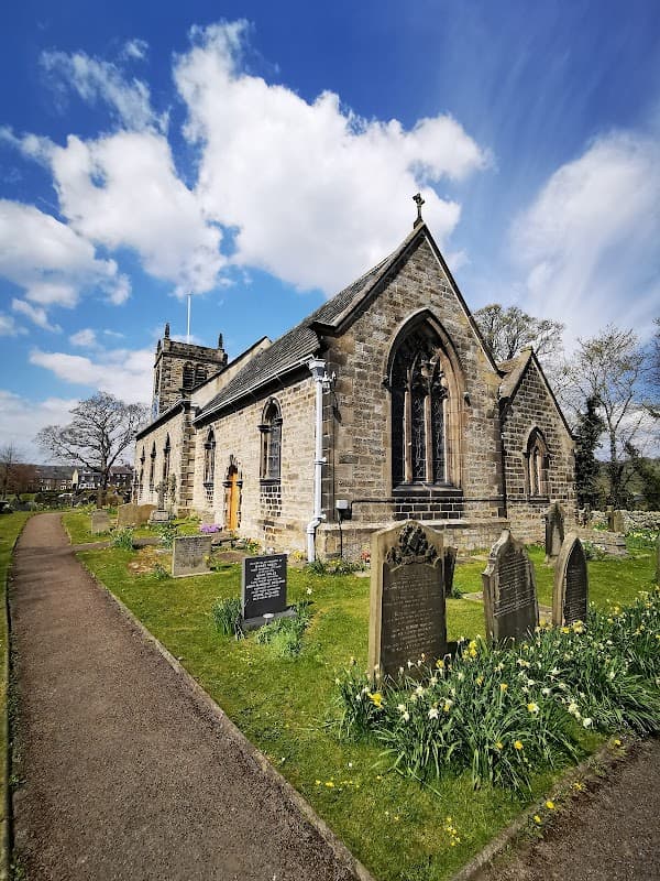St Peter's Church in Addingham, surrounded by gravestones and blooming flowers under a blue sky with clouds.